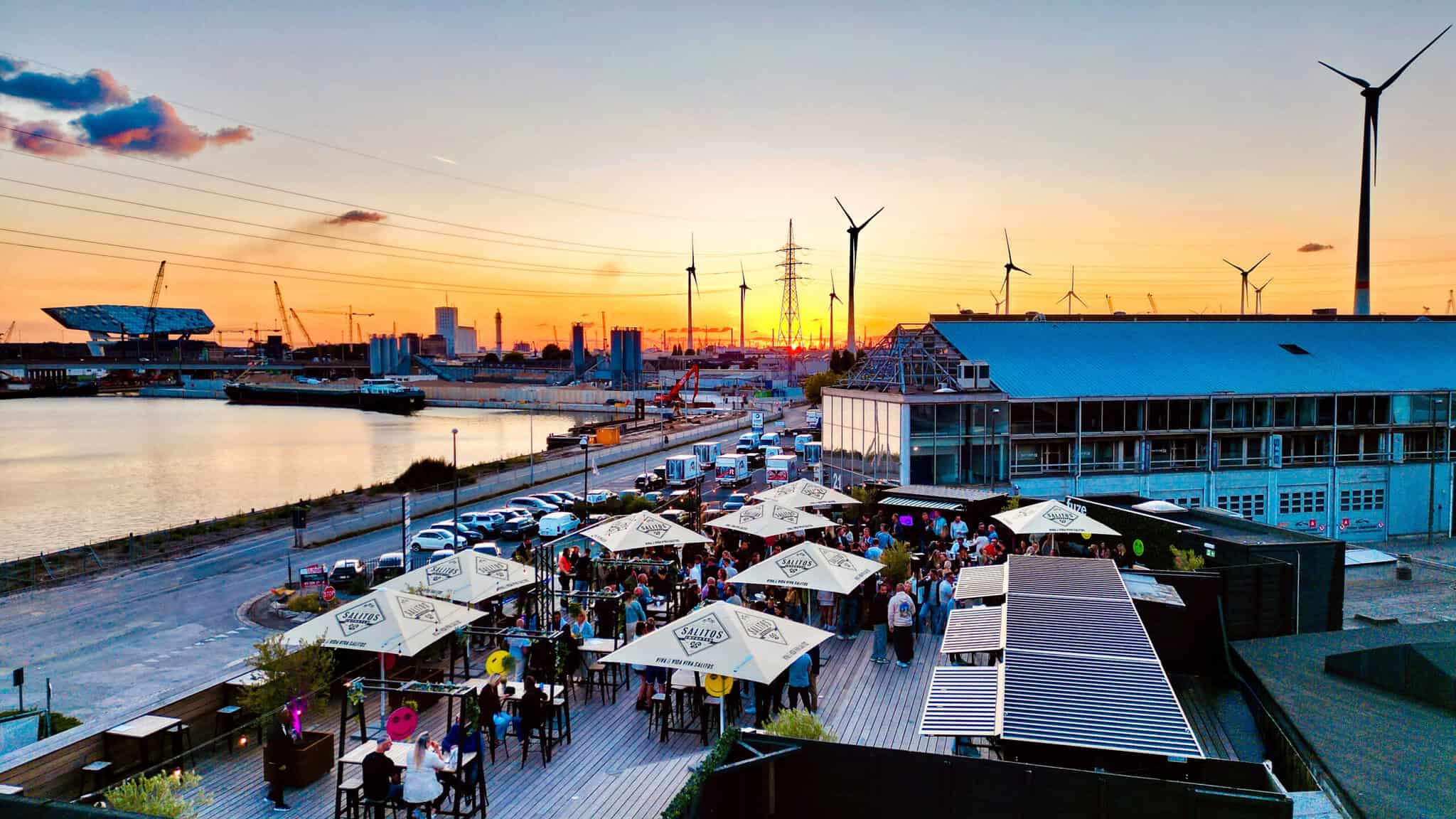 Gezellige zomermarkt aan de Scheldekaaien in Antwerpen met kraampjes, bezoekers en windmolens op de achtergrond tijdens zonsondergang.