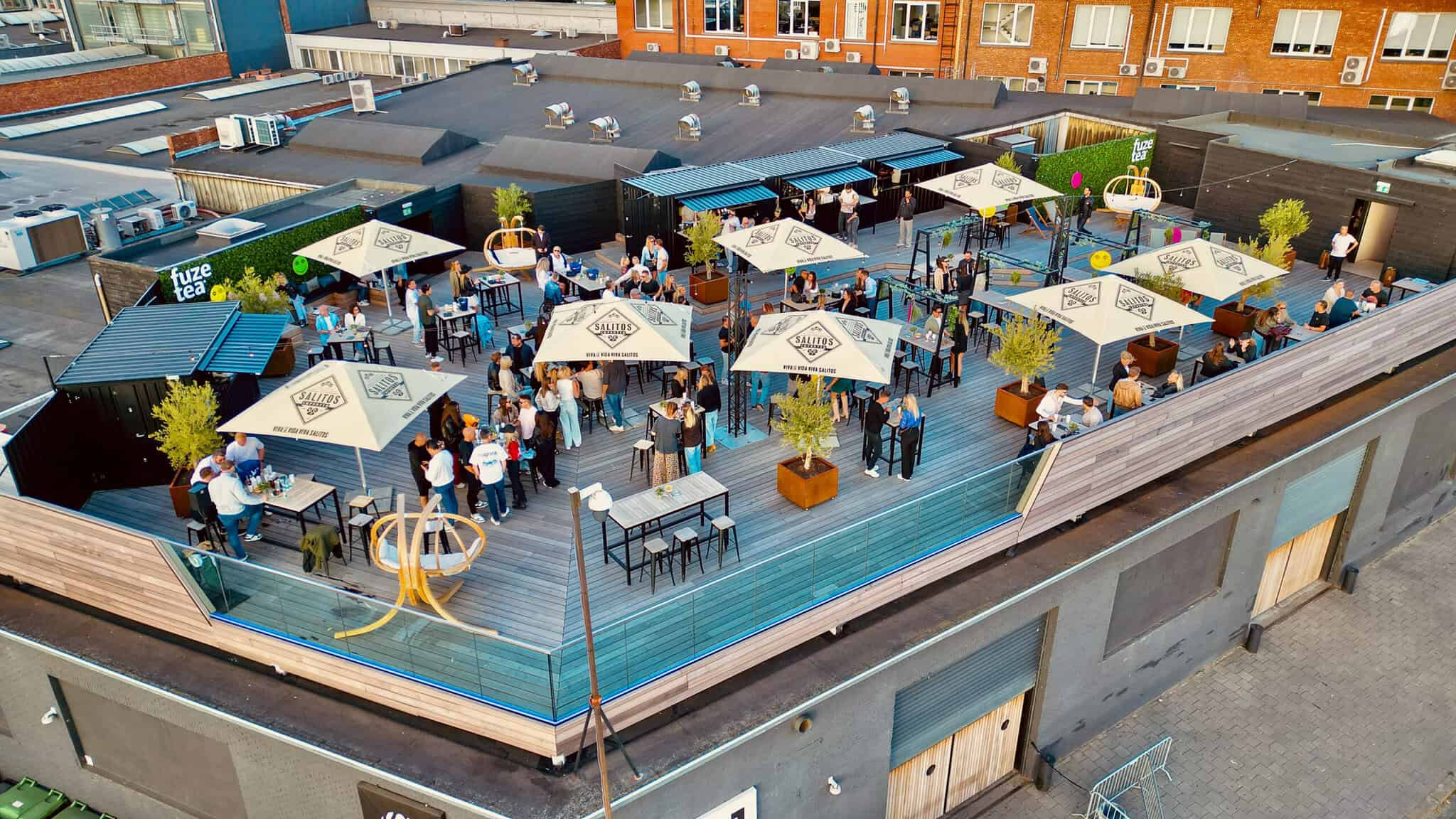 Bovenaanzicht van een levendige rooftop bar in Antwerpen met terras, parasols, en gasten die genieten van een drankje en sociale sfeer.